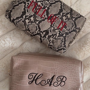Puede incluir: Dos neceseres rectangulares. El neceser superior presenta un patr&oacute;n de piel de serpiente gris y negro con el texto bordado en rojo FULL OF IT. El neceser inferior es de color beige liso con textura de piel de cocodrilo y el monograma negro HAB.