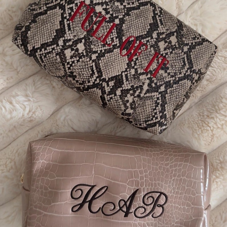 Puede incluir: Dos neceseres rectangulares. El neceser superior presenta un patr&oacute;n de piel de serpiente gris y negro con el texto bordado en rojo FULL OF IT. El neceser inferior es de color beige liso con textura de piel de cocodrilo y el monograma negro HAB.