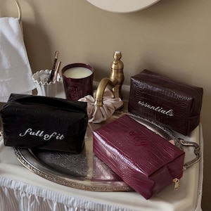 May include: Three rectangular cosmetic bags with a crocodile-embossed texture. One black bag features the white text "full of it," one dark brown bag displays the white text "essentials," and one burgundy bag has the word "Ally" embossed on the front.