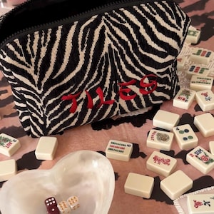 May include: A black and white zebra-striped zippered pouch with the word "TILES" embroidered in red. The pouch is surrounded by ivory-colored mahjong tiles, a heart-shaped dish, and dice.