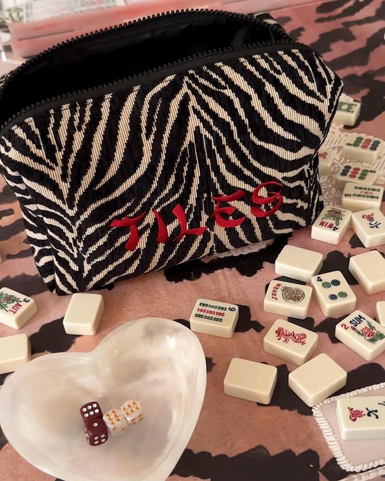 May include: A black and white zebra-striped zippered pouch with the word "TILES" embroidered in red. The pouch is surrounded by ivory-colored mahjong tiles, a heart-shaped dish, and dice.