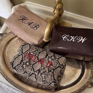 Puede incluir: Tres estuches rectangulares con cremallera y superficies texturizadas. Uno es beige con letras negras bordadas HAB, otro es marr&oacute;n oscuro con letras blancas bordadas CKW, y otro presenta un patr&oacute;n de piel de serpiente con el texto rojo bordado FULL OF IT.