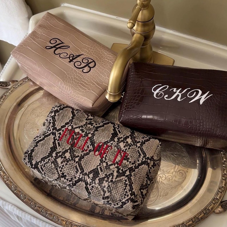 Puede incluir: Tres estuches rectangulares con cremallera y superficies texturizadas. Uno es beige con letras negras bordadas HAB, otro es marr&oacute;n oscuro con letras blancas bordadas CKW, y otro presenta un patr&oacute;n de piel de serpiente con el texto rojo bordado FULL OF IT.