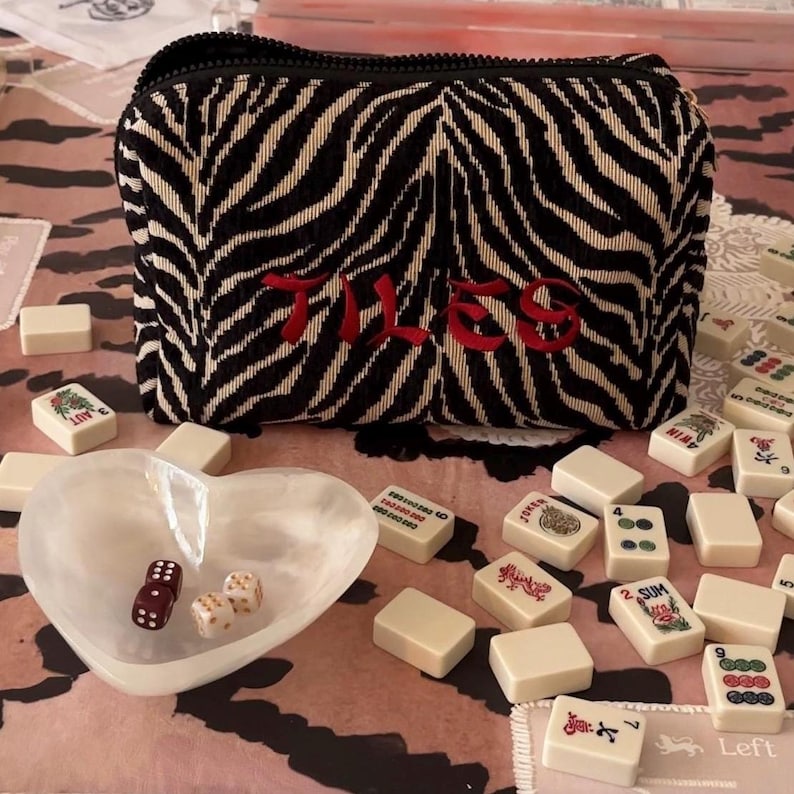 May include: A black and white zebra-striped pouch with the word "TILES" embroidered in red. The pouch is surrounded by white mahjong tiles, a heart-shaped dish, and dice.