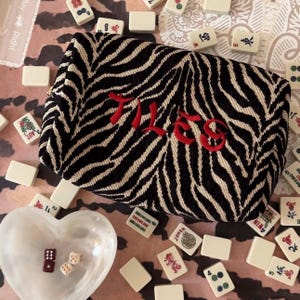 May include: A black and white zebra-striped pouch with the word "TILES" embroidered in red. The pouch is surrounded by ivory-colored mahjong tiles, dice, and a heart-shaped dish. The game pieces are scattered on a patterned surface.