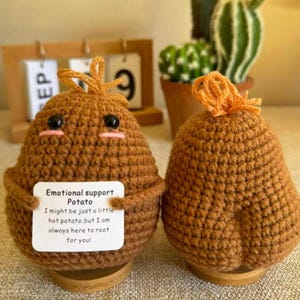 May include: Two handmade crochet potato figures. One holds a sign that reads "Emotional support Potato. I might be just a little hot potato, but I am always here to root for you!" The potatoes are brown with black eyes and orange accents.