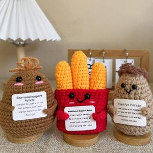May include: Three crocheted potato plushies holding signs with encouraging messages. The first potato is brown and says "Emotional support potato". The second potato is red and yellow and says "Emotional support fries". The third potato is brown and says "Positive potato".