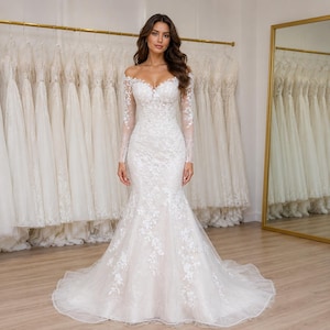 May include: A floor-length white wedding dress with an off-the-shoulder neckline and long sheer sleeves. The gown features a fitted bodice and a flared mermaid skirt, both adorned with intricate white floral lace appliques over a light fabric.