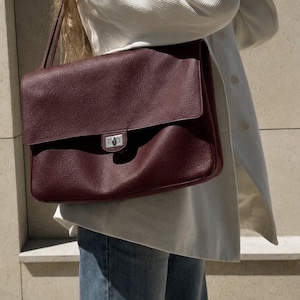 May include: A deep burgundy, rectangular shoulder bag with a textured, pebbled surface. The bag features a large front flap that covers the main compartment and is secured with a silver-toned metal turn-lock closure centered on the front panel.