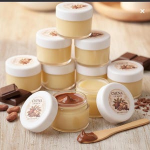 May include: A selection of small, clear lip balm jars with white lids, some arranged in a stack. One jar is open, displaying a chocolate-coloured balm. The lids are labelled with "CHENA LIP BALM" and a cocoa design. Chocolate pieces and cocoa beans are scattered around.