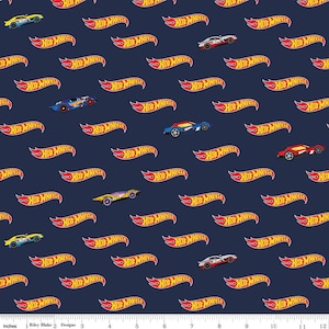 May include: A navy blue fabric with a repeating pattern of red and yellow "Hot Wheels" logos and colorful toy cars. The fabric is 12 inches wide and has a ruler marked in inches along the bottom.
