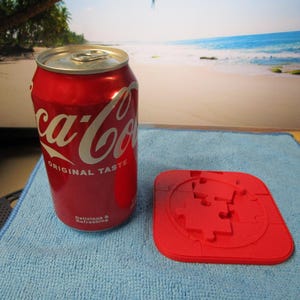 May include: A red Coca-Cola can with white text and a silver lid sits next to a red puzzle. The can reads "Original Taste" and "Delicious & Refreshing". The puzzle is a square shape with a circular design.