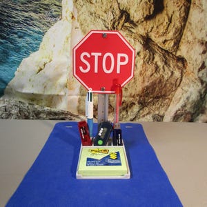 May include: A novelty desk organizer featuring a red stop sign with white text. The organizer holds pens, USB drives, and a Post-it note pad. The base is white, and the background is a blue surface.