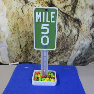 May include: A green and white mile marker sign that reads "MILE 50". The sign is mounted on a silver post and sits in a white tray filled with colorful candies. The background is a textured, neutral color.
