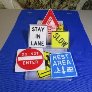 May include: A collection of miniature road signs, including "STAY IN LANE", "DO NOT ENTER", and "REST AREA". The signs are in various shapes and colors, with white, red, yellow, and blue backgrounds. The signs are arranged on a blue surface.