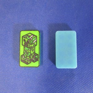 May include: Two rectangular game pieces. One is green with a black and green pixelated Creeper design. The other is a solid light blue color. Both pieces are on a blue textured surface.