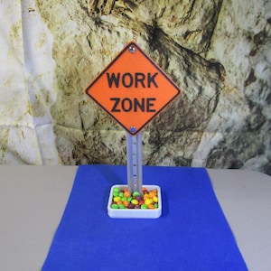 May include: An orange and black "WORK ZONE" road sign with a white base filled with colorful candies. The sign is mounted on a gray post, set against a blue surface and a textured background.