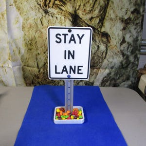 May include: A white sign with black text that reads "STAY IN LANE" is mounted on a gray post. The sign is above a small white tray filled with colorful candies. The background is a blue surface and a textured wall.