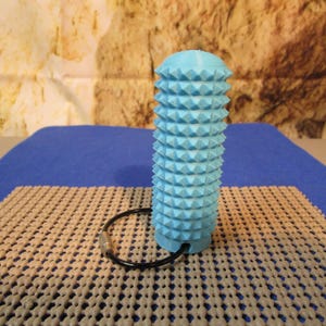 May include: A light blue, cylindrical massage tool with a textured, studded surface. The tool has a black ring attached to its base. The tool is on a beige and blue surface, with a textured background.