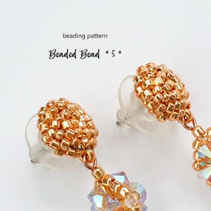 Pode incluir: Um par de brincos de contas douradas com uma gota de cristal. Os brincos são feitos com pequenas contas redondas e têm um design simples e elegante. Os brincos são chamados de "Beaded Bead" e fazem parte de um padrão de contas.