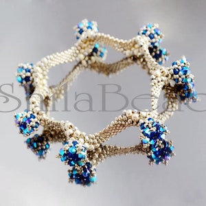 K&ouml;nnte beinhalten: Ein Armband aus silberfarbenen Perlen mit Anh&auml;ngern aus blauen und irisierenden Perlen. Das kreisf&ouml;rmige Design des Armbands spiegelt sich auf einer gl&auml;nzenden Oberfl&auml;che wider. Die filigranen Details unterstreichen die Handwerkskunst des Schmucks.