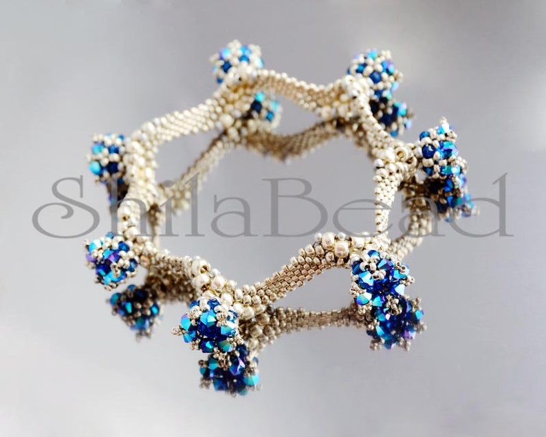 K&ouml;nnte beinhalten: Ein Armband aus silberfarbenen Perlen mit Anh&auml;ngern aus blauen und irisierenden Perlen. Das kreisf&ouml;rmige Design des Armbands spiegelt sich auf einer gl&auml;nzenden Oberfl&auml;che wider. Die filigranen Details unterstreichen die Handwerkskunst des Schmucks.