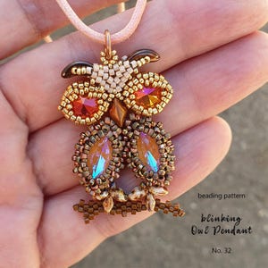 May include: A beaded owl pendant with orange and gold tones, featuring large, iridescent crystal-like eyes. The owl is crafted with small beads and has a brown beaded branch. The pendant hangs from a pink cord. Text on the image reads "beading pattern blinking Owl Pendant No. 32."