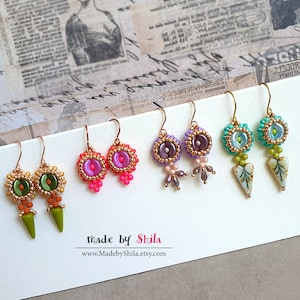 Könnte beinhalten: Vier Paar handgefertigte Ohrringe mit verschiedenen farbigen Perlen und Designs. Die Ohrringe werden auf einer weißen Karte mit dem Text "made by Shila" und einer Website-Adresse präsentiert.