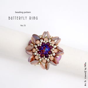 Puede incluir: Un anillo de mariposa con cuentas, con un grupo central de cuentas moradas y azules, rodeado de cuentas plateadas y cuentas en forma de p&eacute;talo en tonos malva y melocot&oacute;n. El anillo se muestra sobre una superficie blanca. El texto dice "Butterfly Ring" y "No 25".