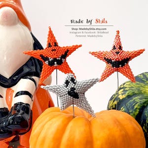Może przedstawiać: Ozdoby w kształcie gwiazd z koralików na Halloween w kolorze pomarańczowym i białym, z czarnymi rysami twarzy. Jedna gwiazda jest biała z czarną twarzą ducha. Ozdoby są wystawione na dyni. Widoczny jest tekst "made by Shila".