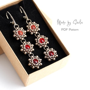 Puede incluir: Pendientes colgantes con centros de piedras preciosas rojas y detalles de cuentas plateadas, presentados en una caja pequeña. Los pendientes tienen un diseño floral y están suspendidos de ganchos plateados. El texto en la caja dice "Made by Shila PDF Pattern."