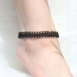 Könnte beinhalten: Ein schwarzes Kettenfußkettchen mit einem zarten, verflochtenen Design. Das Fußkettchen ist aus Metall und wird am Knöchel getragen.