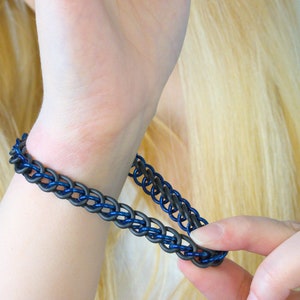 A blue metal and black rubber chain bracelet stretched away from a models wrist with her other hand to show how stretchy it is. These chainmail bracelets stretch about half again their length so you can slip it on over your hand easily.