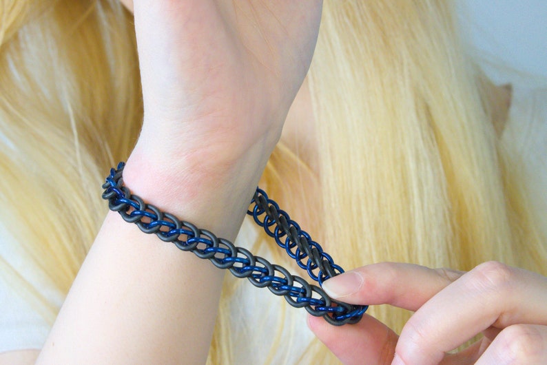 A blue metal and black rubber chain bracelet stretched away from a models wrist with her other hand to show how stretchy it is. These chainmail bracelets stretch about half again their length so you can slip it on over your hand easily.