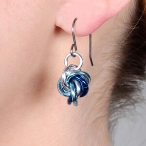 May include: A silver-toned earring with a knot design, featuring interwoven blue and silver metal rings. The earring is suspended from a simple hook, showcasing a blend of cool tones and intricate craftsmanship. The earring is shown on a person's ear.