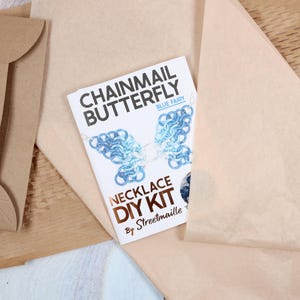 A chainmail butterfly kit sitting on top of the paper packaging it comes wrapped in. The kit is folded into a small tutorial booklet with a small cutout on the cover to show the color of the craft supplies inside the kit. The white paper booklet is highlighted by its contrast against the brown wrapping.