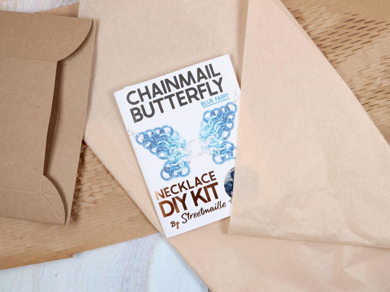 A chainmail butterfly kit sitting on top of the paper packaging it comes wrapped in. The kit is folded into a small tutorial booklet with a small cutout on the cover to show the color of the craft supplies inside the kit. The white paper booklet is highlighted by its contrast against the brown wrapping.