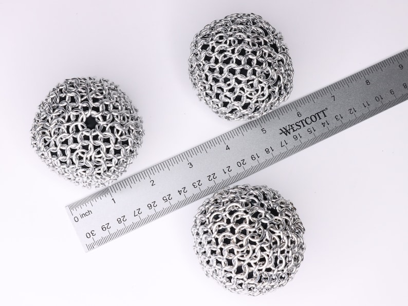 An example of three finished chainmail balls made with silver anodized aluminum rings and filled with black cloth, sitting next to a ruler. Each ball is about 2.5 inches in diameter.