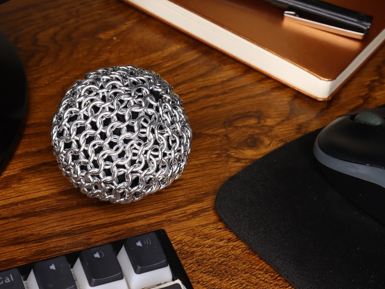 An example of a finished chainmail ball made with silver anodized aluminum rings and filled with black cloth sitting on a wooden desk.