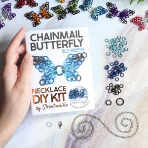 A hand holds a DIY jewelry making kit for a chainmail butterfly necklace. The craft kit is wrapped inside of a folded tutorial booklet. The supplies are laid next to the kit, including a few piles of anodized aluminum jump rings, a plated iron chain, and a plated brass clasp.