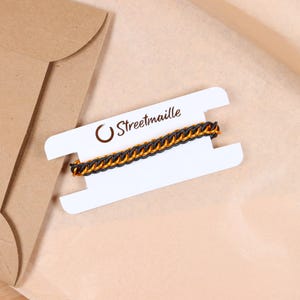 A custom stretchy chain bracelet sitting on top of the paper packaging it comes wrapped in. The bracelet is wrapped around a white paper card to keep it untangled during shipping. The Streetmaille logo is printed on the top of the card in a copper colored foil. The white bracelet card is highlighted by its contrast against the brown wrapping paper and mailer.