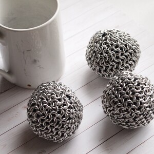 Peut inclure: Une tasse en c&eacute;ramique blanche avec une anse est plac&eacute;e &agrave; c&ocirc;t&eacute; de trois &eacute;ponges en cotte de mailles en acier inoxydable. Les &eacute;ponges sont sph&eacute;riques et compos&eacute;es d'anneaux m&eacute;talliques imbriqu&eacute;s. Les articles sont dispos&eacute;s sur une surface en bois clair.