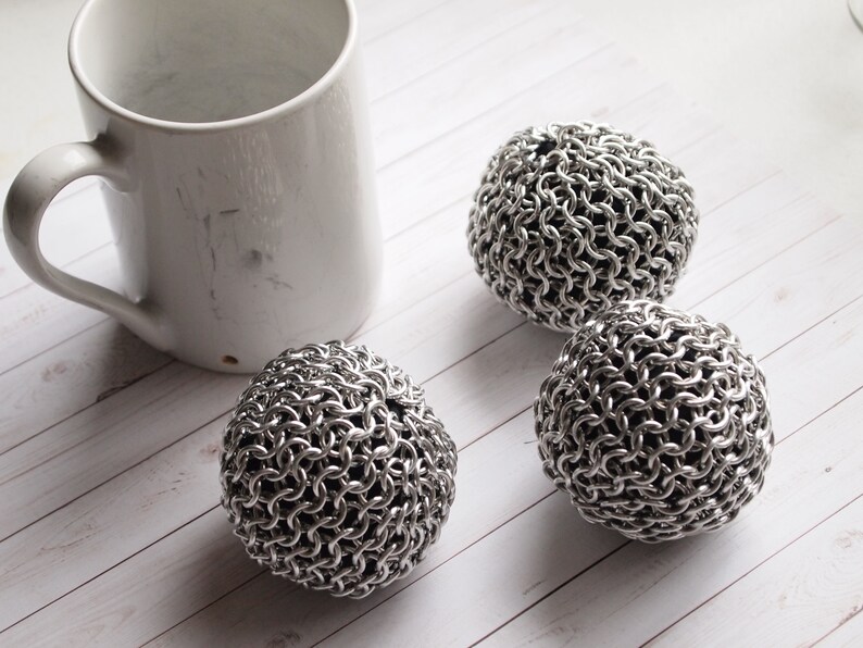 Peut inclure: Une tasse en c&eacute;ramique blanche avec une anse est plac&eacute;e &agrave; c&ocirc;t&eacute; de trois &eacute;ponges en cotte de mailles en acier inoxydable. Les &eacute;ponges sont sph&eacute;riques et compos&eacute;es d'anneaux m&eacute;talliques imbriqu&eacute;s. Les articles sont dispos&eacute;s sur une surface en bois clair.