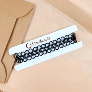 An oversized chainmail tattoo choker sitting on top of the paper packaging it comes wrapped in. The necklace is wrapped around a white paper card to keep it untangled during shipping. The Streetmaille logo is printed on the top of the card in a copper colored foil. The white necklace card is highlighted by its contrast against the brown wrapping paper and mailer.