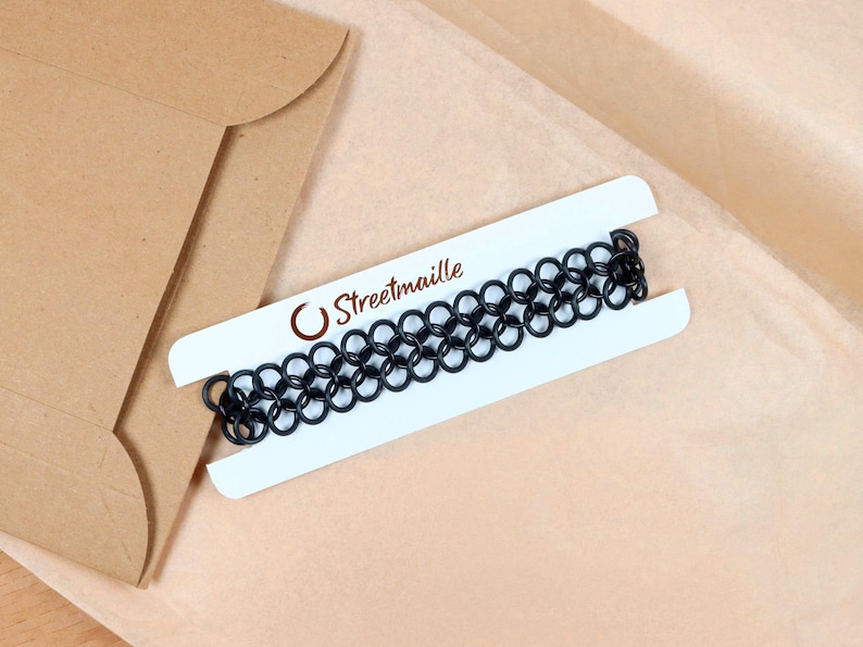 An oversized chainmail tattoo choker sitting on top of the paper packaging it comes wrapped in. The necklace is wrapped around a white paper card to keep it untangled during shipping. The Streetmaille logo is printed on the top of the card in a copper colored foil. The white necklace card is highlighted by its contrast against the brown wrapping paper and mailer.