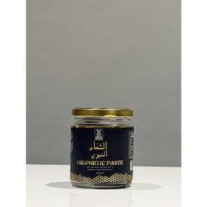 Op de afbeelding: Een glazen pot met een gouden deksel en een zwart etiket. Het etiket bevat Arabische tekst en de woorden "Prophetic Paste". De pot bevat een donkere, viskeuze substantie. De achtergrond is neutraal grijs.