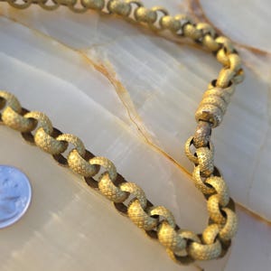 May include: A gold-toned chain necklace with textured links. The necklace is displayed on a light-colored surface. The chain's links are connected in a classic pattern, and the clasp is visible. The necklace is a vintage piece.