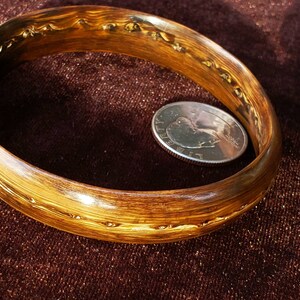 May include: A wooden bangle with a brown and gold finish. The bangle is smooth and polished, with a subtle grain pattern. A US quarter is placed next to the bangle for size comparison.