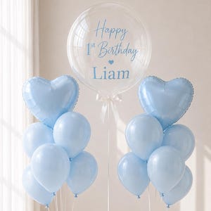 May include: A balloon arrangement featuring a large clear bubble balloon with the blue text Happy 1st Birthday Liam and a small heart icon. It is flanked by two clusters of light blue latex balloons and heart-shaped foil balloons on white ribbons.