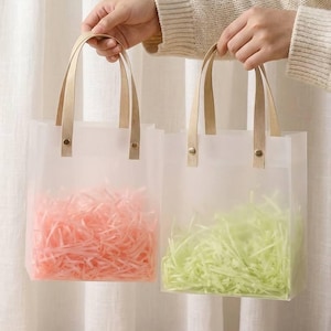 May include: Two clear gift bags with tan handles, each filled with colorful paper shred. One bag contains pink shred, the other green. The bags are held up against a white curtain background.